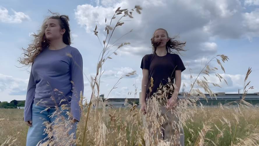 The field – Zwei Personen stehen auf einer Wiese mit hohem Gras. Beide haben lange Haare. Der Himmel ist bewölkt. Im Hintergrund ist ein Gebäude zu sehen.