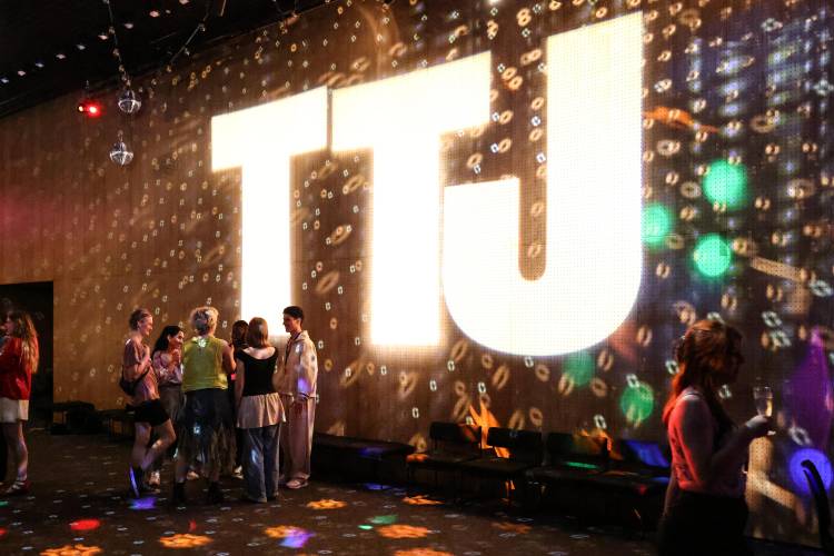 Theatertreffen der Jugend – In einem abgedunkelten Raum wird ein großes TTJ-Logo an die Wand und Lichter an die Wand projeziert. Davor stehen junge Leute in Gruppen.