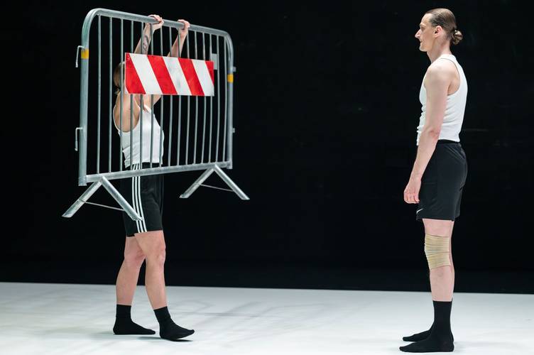 Tanz im August: CLAP & SLAP 1 – Zwei Performer*innen vor schwarzem Hintergrund, eine hält einen Absperrzaun in die Höhe.