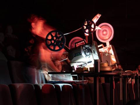 Warm Data – Filmprojektor mit rotierenden Filmspulen in einem dunklen Raum auf klappbaren Kinositzen. Verschwommenes Abbild einer Person daneben.