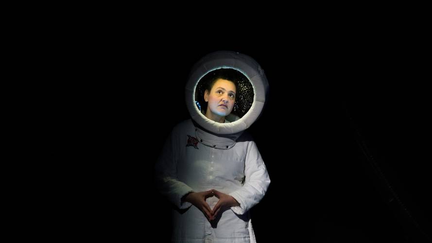Person in Astronautenkostüm im Dunkeln, beleuchtetes Gesicht im Helm, Hände vor dem Körper gefaltet, nachdenklicher Blick nach oben gerichtet.