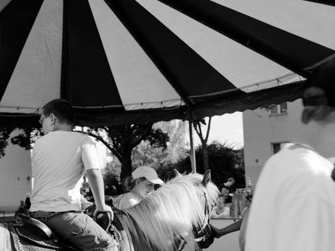 Akinbode Akinbiyi, Aus der Serie: African Quarter, seit den 1990er Jahren – Schwarz-Weiß-Fotografie: Seitenansicht von einem Jungen auf einem Pferd unter einem gestreiften Baldachin. Im Hintergrund sind weitere Menschen zu sehen.