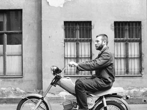 Entwurfsmodell Mokick „Simson S 50“ im Hof des Ateliers von Lutz Rudolph am Helmholtzplatz, Prenzlauer Berg, Fotografie, um 1967 Gestaltung: Lutz Rudolph, Karl Clauss Dietel, Privatarchiv Ania Rudolph – Fotografie eines Mannes sitzend auf einem Motorrad-Modell in einem Berliner Hinterhof