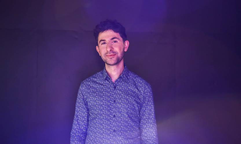 Alex Raineri – Person mit lockigem Haar, gemustertem Hemd vor dunklem Hintergrund, in lila Licht beleuchtet, lächelt leicht in die Kamera.