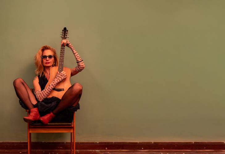 Person mit Sonnenbrille sitzt auf einem Stuhl, hält eine Gitarre, trägt gemusterte Armstulpen, schwarze Strümpfe und rote Stiefel vor grüner Wand.