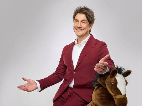 Matze Knop - Spitzenreiter – Eine Person in einem weinroten Anzug sitzt lächelnd auf einem braunen Spielzeug-Pferd vor grauem Hintergrund.