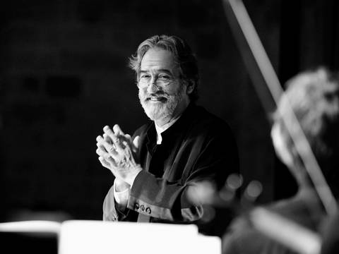 Jordi Savall – Ein älterer Mann mit Bart hält seine Hände vor der Brust verschränkt und lacht in die Kamera. Er steht vor einem Dirigentenpult an der Seite sind unscharf Streichinstrumente zu erkennen.