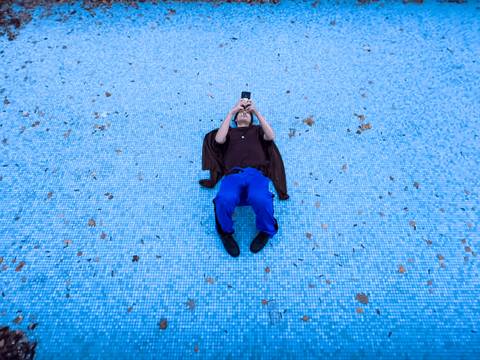  – Person liegt auf dem Rücken auf blauem Boden, trägt blaue Hose, schwarzen Pulli, schwarze Schuhe und hält ein Smartphone nach oben gerichtet.