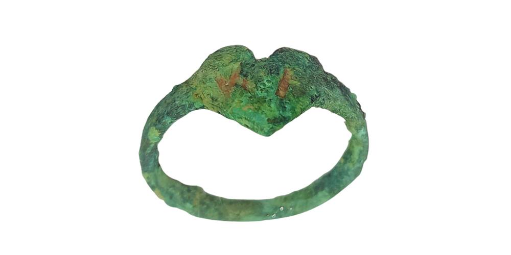 Kupferner Ring mit einem Herz und Initialen aus dem Zwangsarbeitslager Berlin-Köpenick – Ein schmaler, grünlich oxidierter Ring mit unregelmäßiger Oberfläche. Die Ringschiene ist dünn und wirkt stellenweise uneben. Auf der Vorderseite befindet sich eine herzförmige Platte mit zwei eingravierten, senkrechten Linien. Die Patina deutet auf ein altes oder antikes Schmuckstück hin.