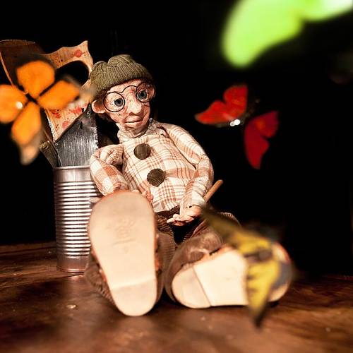 Puppe mit Brille und Mütze sitzt auf Holzboden. Schmetterlinge fliegen umher. Konservendose mit bemaltem Holz dahinter.