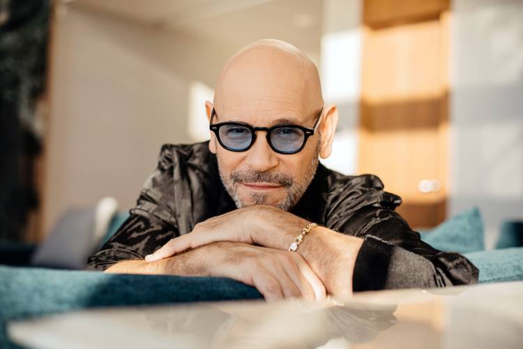Rami Kleinstein – Männlich gelesene Person mit Glatze und Bart, trägt dunkle Sonnenbrille und schwarze Jacke, stützt sich auf Arme, lächelt leicht, Hintergrund unscharf.