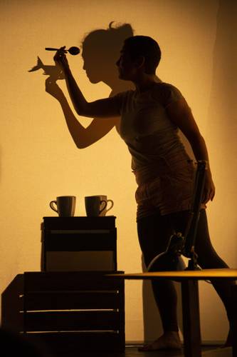 Schattenwerfer – Person hält Löffel vor Lichtquelle, projiziert Schattenszene mit Vogel auf Wand. Zwei Tassen auf Regal, starke Licht-Schatten-Kontraste.