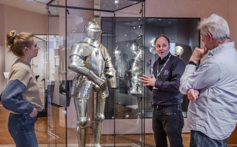 Ein Museumsführer steht vor einer Ritterrüstung in einer Vitrine und erklärt sie zwei Personen.