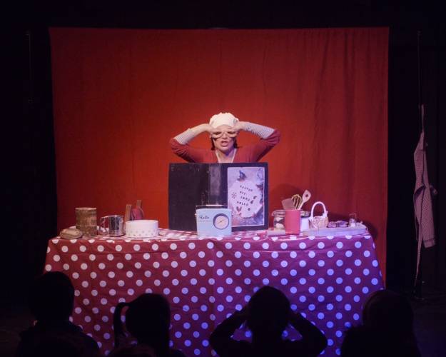 Person mit weißer Mütze hinter Tisch mit roter Tupfentischdecke, Backutensilien und Schild "Backen mit Frau Holle", roter Hintergrund, Publikum im Vordergrund.