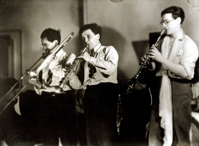 Fotograf*in: Unbekannt, Die Bauhaus-Kapelle, mit Hermann Clemens Röseler, Xanti Schawinsky und T. Lux Feininger, 1925 – Drei Männer in dunklen Hosen und hellen Hemden geben mit ihren Blasinstrumenten ein Konzert. Der Mann links spielt Posaune, der Mann in der Mitte Klarnette und der Mann rechts Oboe.