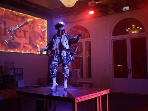 Gira – Portraitperformance Natasha Vergilio – Person in bunter Kleidung und Perücke tanzt auf einem Tisch, im Hintergrund Projektion mit Text "her" und Händen über Flammen. Raum mit roten Lichtern und