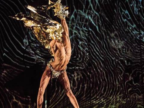 Queer Performance Festival – Person wirbelt mit goldglänzendem Stoff vor dunklem, wellenförmigem Hintergrund.