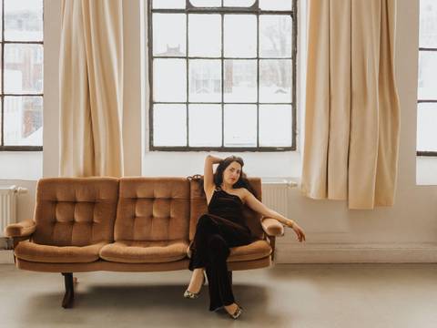 Anna Margolina – Anna Margolina sitzt in einem lofartigen Raum mit großen Fenstern und cremefarbenen Vorhängen auf einem braunen Vintage-Velour-Sofa. Sie trägt ein schwarzes Kleid und Pumps.