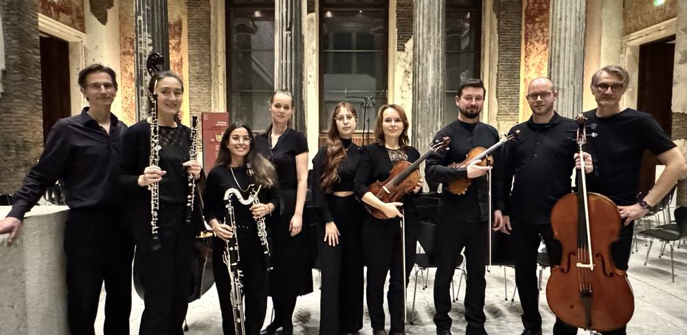 Ensemble MEMORIA TENERE – Acht Musiker*innen mit Instrumenten in dunkler Kleidung stehen vor antik wirkenden Saeulen in einem Saal. Links Klarinetten, rechts Geigen und Cello.