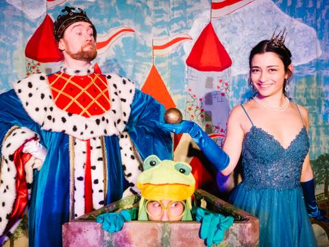  – Eine Person in einem königlichen Umhang, ein großer Frosch mit Brille und eine lächelnde Person in einem blauen Kleid mit Krone posieren vor einer Kulisse.