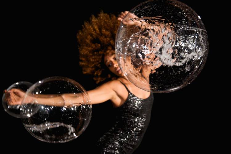 Brazilian urban Dances - Passinho und Rebolado 1 – Eine Performerin in schwarzen Kleid in Bewegung, vor ihr zwei Seifenblasen.