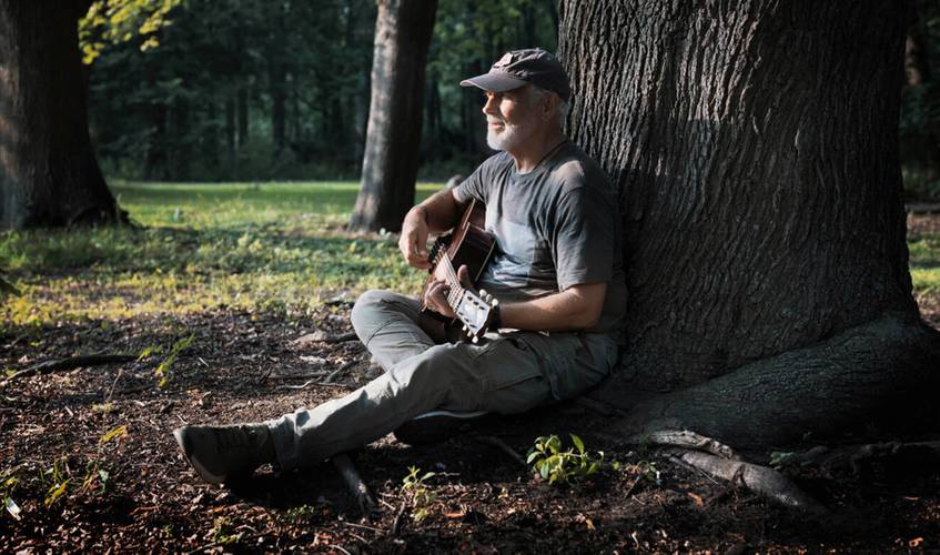 Person mit grauem Bart, dunklem T-Shirt und Cap sitzt im Wald mit Gitarre an einen Baum gelehnt. Sonnenlicht fällt durch das Blätterdach.