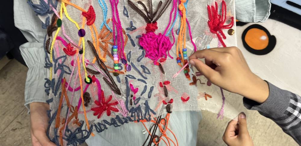 Zwei Hände sticken bunte Muster aus Garn und Perlen auf transparentem Stoff. Person trägt blaugraues Outfit. Stickerei mit abstrakten Formen.