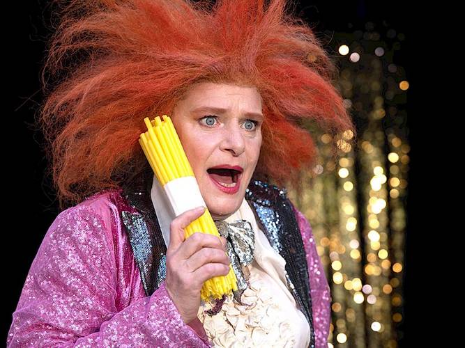 Märchen im Glaspalast von und mit dem Hexenberg Ensemble: Das Mädchen mit den Schwefelhölzern I Rumpelstilzchen – Person mit orange-rotem, vollem Haar hält gelbe Hartweizennudeln ans Ohr. Glitzerndes, pinkfarbenes Outfit mit großer Schleife. Überraschter Gesichtsausdruck.