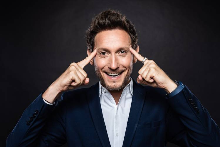 LiorSuchard – Person mit bart lächelt, zeigt mit zeigefingern auf die stirn. Trägt dunkelblauen anzug und weißes hemd. Hintergrund dunkel.