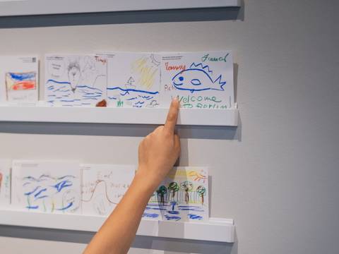 Zeichnungen im Humboldt Labor – Eine Hand zeigt auf bunte Zeichnungen an einer Wand, darunter ein Fisch mit dem Schriftzug "Willkommen in Berlin!".