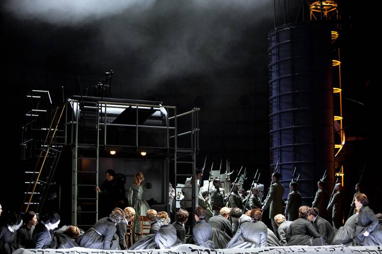 Nabucco – Bühnenbild mit vielen Darsteller*innen in historischem Militär-Outfit vor einem großen Zylinder und einer Metallkonstruktion mit Treppe. Rauch im Hintergrund.