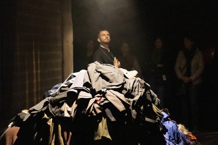 Stapel bunter Kleidung im Vordergrund, dahinter steht eine männlich gelesene Person im Scheinwerferlicht, umgeben von mehreren Personen im Hintergrund.