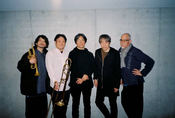Otomo Yoshihide New Jazz Quintet – Fünf Personen stehen vor einer grauen Wand; zwei halten Instrumente, eine Trompete und eine Posaune.