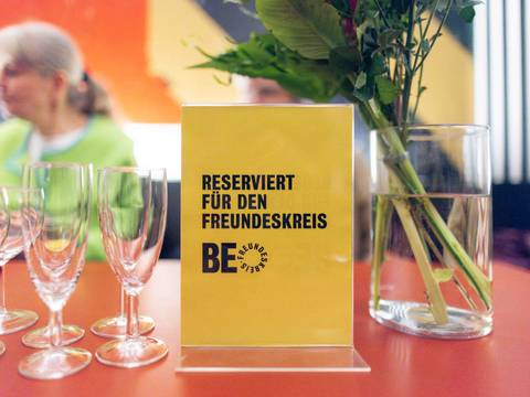 Freundeskreis – Gelbes Schild mit Text "RESERVIERT FÜR DEN FREUNDESKREIS BE". Daneben Blumen in Glasvase, mehrere leere Weingläser, unscharfe Person im Hintergrund.