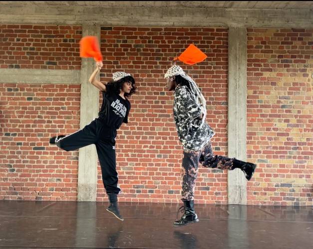 UNEXPECTED LESSON 2: Dabke-Workshop 1 – Zwei Personen in einer Tanzbewegung vor einer Backsteinwand. Sie halten orangene Fahnen in der Hand.