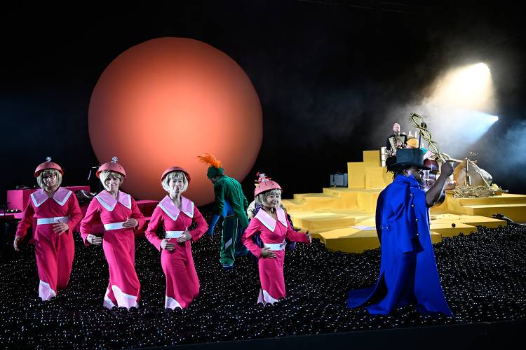 Sunville – Fünf Personen in pinken Outfits mit Hüten auf schwarzem Boden, gelbe Stufen, große rote Scheibe im Hintergrund, jemand im blauen Umhang und schwarzem Hut.