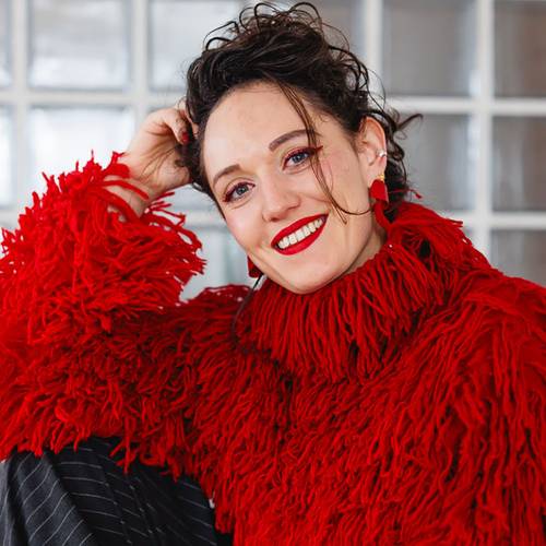 Elena Artisi – Person mit rotem Strickpullover und rotem Lippenstift lächelt in die Kamera. Hintergrund aus Glasblöcken.