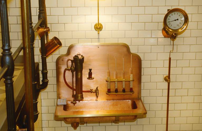 Am Spindeltisch wird mit Würzspindeln der Malzzuckergehalt bestimmt und so der Gärprozess kontrolliert. – Waschbeckenartiger Aufbau aus Kupfer an einer weiß gekachelten Wand.