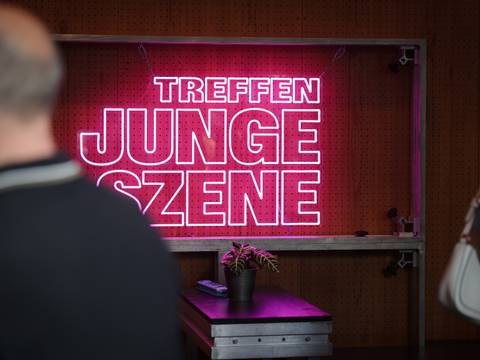  – An der Wand ist ein leuchtender, pinkfarbener Schriftzug mit der Aufschrift „Treffen junge Szene“ zu sehen, der in dem ansonsten dunklen Raum besonders hervorsticht.