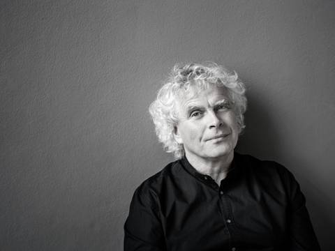 Sir Simon Rattle – Portrait eines Mannes mit grauen lockigen Haaren und schwarzem Hemd vor einem grauen Hintergrund.