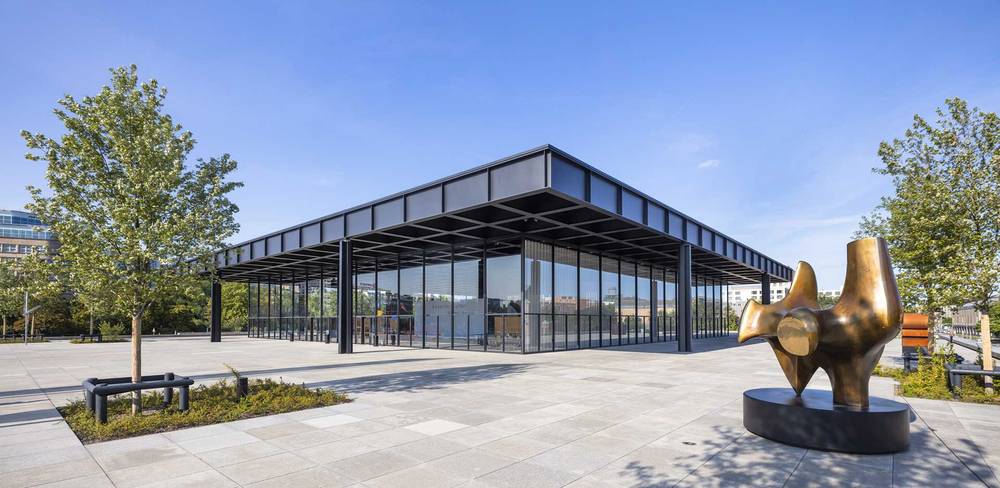 Neue Nationalgalerie – Moderne Glas-Stahl-Gebäudestruktur auf gepflastertem Platz mit abstrakter Bronzeskulptur rechts, umgeben von Bäumen und blauen Himmel.