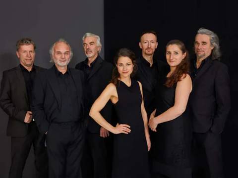 L'ART DE PASSAGE - Vom Tango zum Jazz und zurück – Sieben Personen in schwarzer Kleidung stehen vor dunklem Hintergrund, sechs männlich gelesene und eine weiblich gelesene Person in der Mitte, alle schauen zur