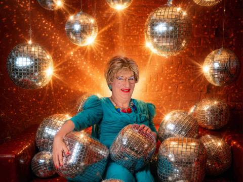 Edith Schröder – Person in blauem Kleid sitzt auf rotem Sofa, umgeben von glitzernden Discokugeln, die von der Decke hängen und auf dem Boden liegen.