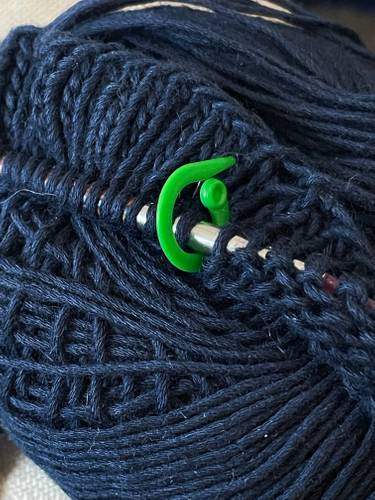 Garn mit Stricknadel und Maschenmarkierer – Zu sehen ist ein dunkelblaues Garnknäuel, in dem eine metallene Stricknadel mit aufgezogenen Maschen steckt. Zwischen zwei Maschen befindet sich ein hellgrüner Maschenmarkierer.