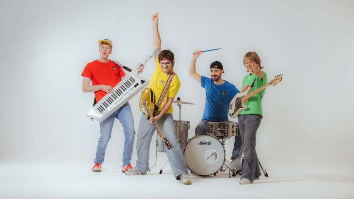 Das große Kinderrechte-Konzert – Vier Personen mit Instrumenten: Gitarre, Keyboard, Schlagzeug, Bass. Farbenfrohe Kleidung in Rot, Gelb, Blau, Gruen. Weisser Hintergrund, energiegeladene Posen.