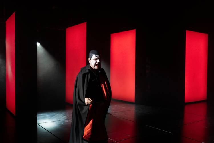 Mephisto – Ein Mann mit weißer Gesichtsschminke und einem rot-schwarzen Umgang steht in einem rot-schwarzem Raum