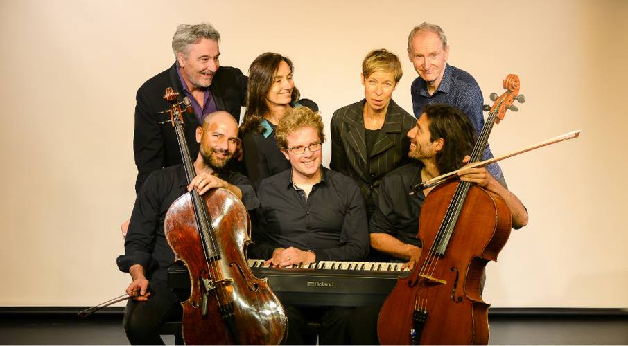 Sieben Personen mit Instrumenten; zwei Cellist*innen im Vordergrund, eine Person am Roland-Klavier, die restlichen im Hintergrund stehend, lächelnd.