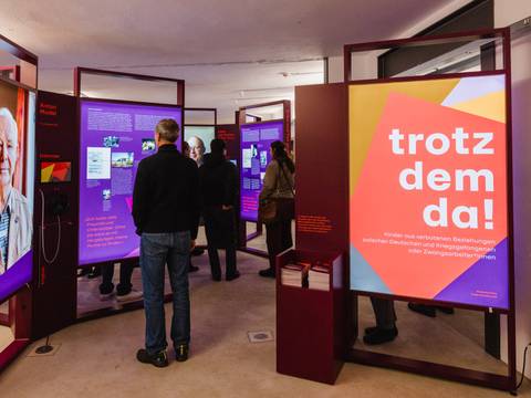  – Mehrere Personen betrachten Ausstellungstafeln in einem Raum, eine Tafel zeigt den Schriftzug 'trotzdem da!' in Weiß auf rotem und gelbem Hintergrund.