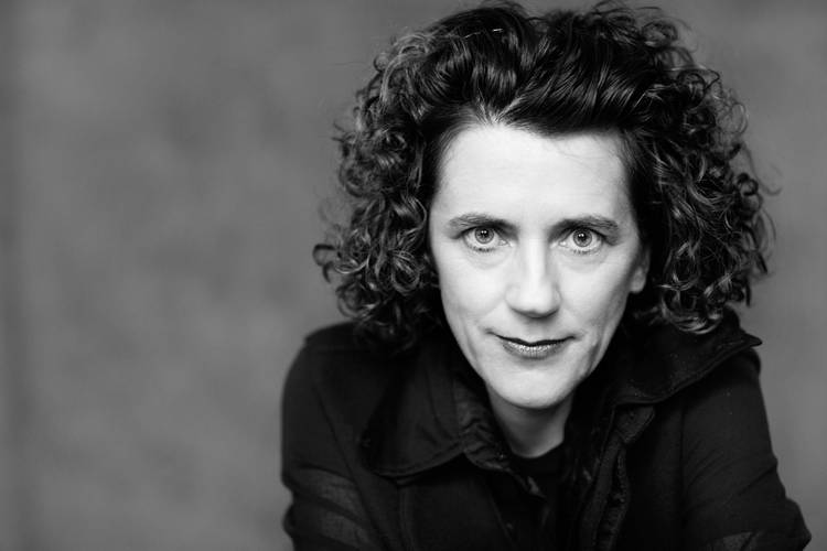 Die Komponistin Olga Neuwirth – Eine Frau mit dunklen lockigen Haaren schaut sehr direkt mit einem Lächeln auf den Lippen in die Kamera.