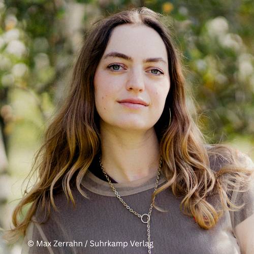 Dana Vowinckel – Person mit langen braunen Haaren und Kette steht draußen vor unscharfem Hintergrund. Text im Bild: © Max Zerrahn / Suhrkamp Verlag.